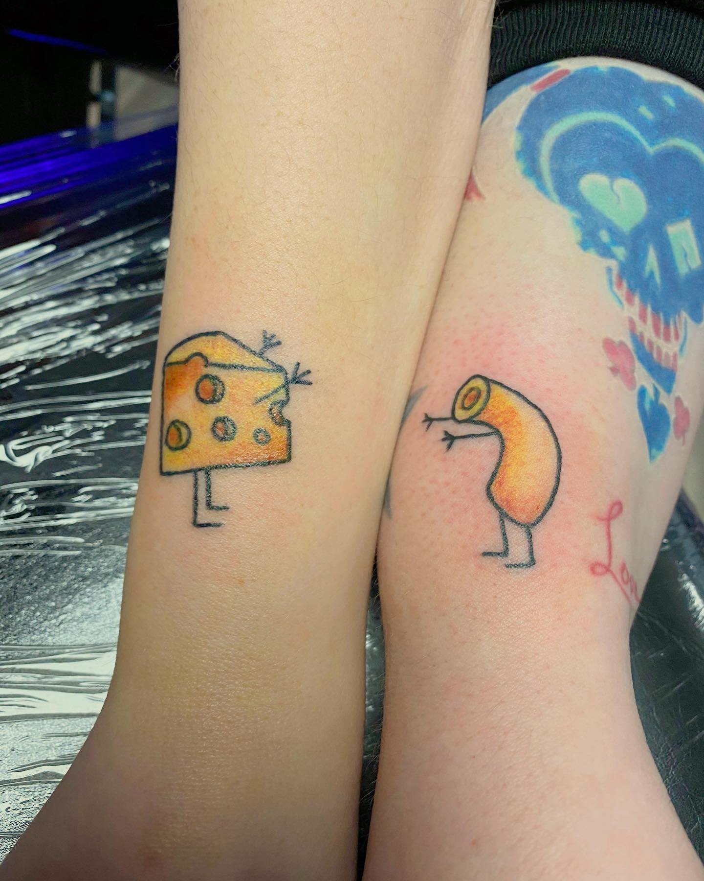 Cheese and macaroni tattoos on legs about to hug each other against a colorful background tattoo
